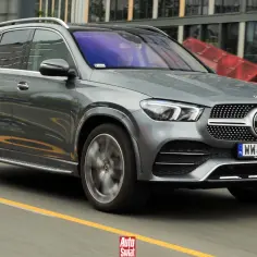 Szary Mercedes GLE 300d jedzie po drodze, za nim nowoczesne budynki.