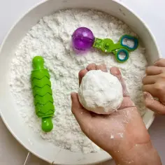 Dziecko bawi się masą sensoryczną, tworząc kulki. W misce są zabawki: wałek i nożyczki. Proste masy sensoryczne dla dzieci przepisy.