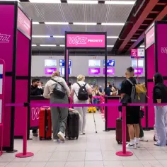Klienci Wizz Air stoją w kolejce do odprawy. Zastanawiasz się, czy można przewozić szkło w bagażu podręcznym Wizzair? Sprawdź wymiary walizek na tablicy.