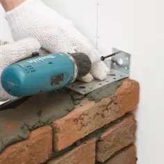 Rękawice i wiertarka pomagają w murowaniu ścianek działowych, mocując metalowy kątownik do ściany.