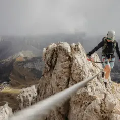 Kobieta w kasku i sprzęcie wspinaczkowym pokonuje via ferrata polska. W tle górzysty krajobraz z krętymi drogami.
