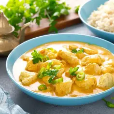 Kurczak w kremowym sosie curry, udekorowany zieloną papryczką i natką pietruszki, obok ryżu.