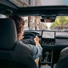 Wnętrze toyoty corolla 2.0 hybrid. Kierowca zapatrzony w nawigację, jedzie ulicą pełną zieleni.