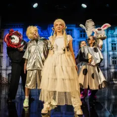Teatr bajka repertuar dla dzieci: postacie w fantazyjnych strojach, w tym królik i dziewczynka w białej sukni, na tle scenografii.