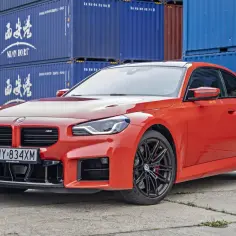 Pomarańczowe BMW M2 Competition z czarnymi felgami stoi na tle niebieskich kontenerów.