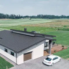 Nowoczesny dom z ciemnym, eleganckim dachem. To najtańsze pokrycie dachowe, które doskonale komponuje się z otaczającą zielenią i białymi ścianami.