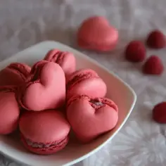 Różowe makaroniki francuskie w kształcie serc na białym talerzyku, obok kilka malin.