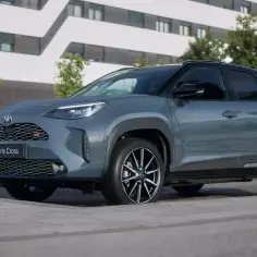 Szary Toyota Yaris Cross benzyna GR Sport z czarnymi dodatkami stoi na chodniku przed nowoczesnym budynkiem.