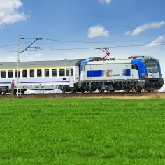 Nowoczesny pociąg TLK PKP Intercity z niebiesko-białą lokomotywą mknie przez zielone pola pod błękitnym niebem.