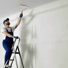 Mężczyzna w niebieskim kombinezonie stoi na drabinie, malując sufit wałkiem.
