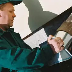 Mężczyzna w zielonej kurtce i czapce montuje rurę, która posłuży do doprowadzenia powietrza do kominka.