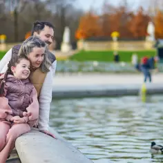 Rodzina cieszy się wspólnym spacerem nad wodą, jakby spędzali wakacje na Mazurach z dziećmi. Widok kaczek pływających w stawie dodaje uroku.