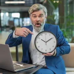 Zaskoczony mężczyzna w garniturze wskazuje na zegar, pytając "deadline co to?". Obok laptop.