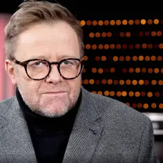 Olaf Lubaszenko w okularach, w szarym garniturze i czarnym golfie, na tle czerwonej kurtyny i świateł.