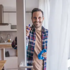 Uśmiechnięty mężczyzna w kratę trzymający butelkę z płynem i niebieską ściereczkę, gotowy do mycia okien.
