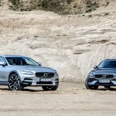 Dwa samochody Volvo Cross Country, srebrny i grafitowy, stoją na piaszczystym podłożu przed piaskową ścianą.