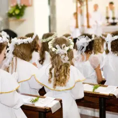 Dzieci w bieli, z wiankami na głowach, siedzą w ławkach kościoła, czekając na moment, kiedy jest komunia święta.