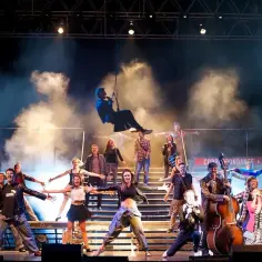 Scena musicalowa z tańczącymi i śpiewającymi ludźmi. W powietrzu wisi mężczyzna na linie, jak w musicalu.