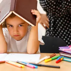 Smutna dziewczynka z książką na głowie, otoczona kredkami i zeszytami. Co jeśli nauczyciel zgubi prace ucznia?
