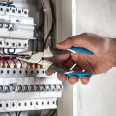 Elektryk pracuje przy rozdzielnicy, dbając o prawidłowy power factor. Widoczne są bezpieczniki i przewody.