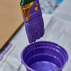 Pędzel zanurzony w fioletowej farbie, z której ściekają krople do plastikowego pojemnika.