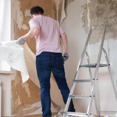 Mężczyzna w rękawiczkach zdejmuje starą tapetę ze ściany, przygotowując pomieszczenie do remontu. W pobliżu stoi drabina.