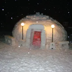 Śnieżne iglo nocą, oświetlone lampami, z czerwonym światłem w środku. Obok ławki ze śniegu.