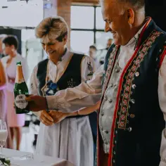 Para w strojach ludowych trzyma butelkę wina musującego na tle weselnych gości.