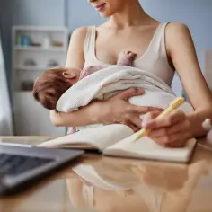 Kobieta trzyma dziecko, pisze w notatniku przy laptopie.