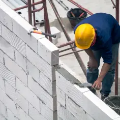 Budowlaniec układa bloczki z betonu komórkowego, dbając o precyzyjne wymiary ściany. Młotek i kielnia czekają na użycie.