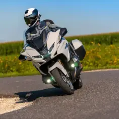 Motocyklista w kasku i stroju podróżuje na białym motocyklu BMW RT 1250 po zakręcie, z polem żółtych kwiatów w tle.