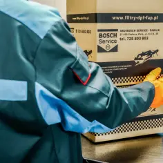 Pracownik w pomarańczowych rękawiczkach trzyma karton z logo Bosch Service. Dowiedz się, ile trwa wypalanie DPF na postoju.