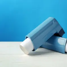 Dwa niebieskie inhalatory na astmę bez recepty leżą na białym blacie na tle błękitnej ściany.