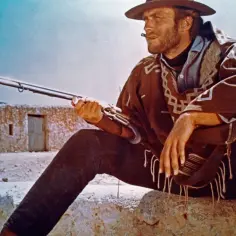 Clint Eastwood w wieku z karabinem, w ponczo i kapeluszu, z papierosem w ustach, siedzi na kamieniu.