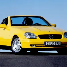 Żółty kabriolet Mercedes SLK 200 z czarnym dachem, zaparkowany na ciemnej drodze pod błękitnym niebem.