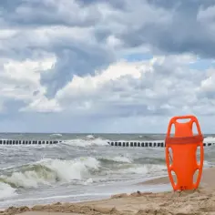 Pomarańczowa deska ratownicza na piaszczystej plaży. W tle wzburzone morze i drewniane pale. Brak kolorów flag, ale ratownik czuwa.
