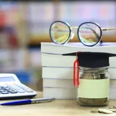 Skarbonka w birecie, stos książek, okulary, kalkulator i długopis. Idealne do zatrudnienia ucznia i studenta do 26 roku życia, brak PIT i ZUS.