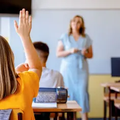 Uczennica z podniesioną ręką na lekcji. Czy po radzie pedagogicznej można zmienić oceny? Nauczycielka w tle.