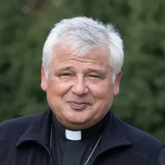 Uśmiechnięty mężczyzna w sutannie i czarnym bezrękawniku, z siwymi włosami. To jałmużnik papieski, który z troską patrzy na świat.