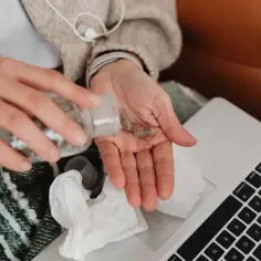 Osoba bierze kapsułki, zastanawiając się, kto płaci za L4 po ustaniu zatrudnienia. Obok leży laptop i chusteczki.