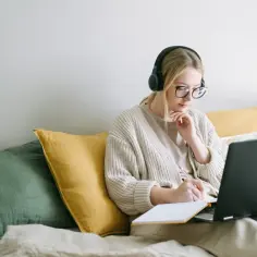 Dziewczyna w słuchawkach i okularach uczy się, jak się pisze opinion essay, notując pomysły w zeszycie przy laptopie.