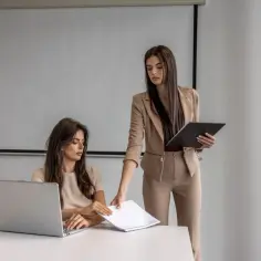 Dwie kobiety w biurze, jedna przy laptopie, druga podaje dokumenty.
