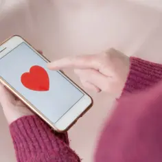 Dłoń z palcem wskazującym na ekranie telefonu, na którym widnieje czerwone serce. To może być ekran startowy aplikacji dla gejów, szukającej miłości.