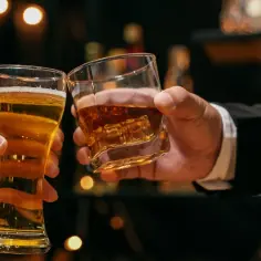 Dwie dłonie unoszą kieliszki: jeden z piwem, drugi z bursztynowym trunkiem. Co lepsze piwo czy wódka? Decyzja należy do Ciebie.