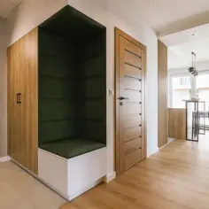Nowoczesny hol z drewnianymi panelami, zieloną wnęką wypoczynkową i drzwiami. W tle widać kuchnię z wysokimi stołkami. Białe listwy podkreślają architekturę.