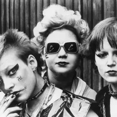Trzy kobiety w stylu punk z charakterystycznym makijażem i fryzurami. Jedna z nich pali papierosa, druga ma okulary przeciwsłoneczne, a trzecia ozdobę w ustach.
