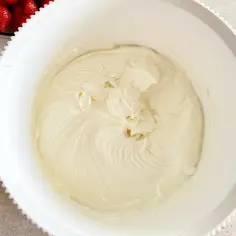 Gładka, kremowa masa z mascarpone w białej misie, gotowa do deseru. Obok miski widać miseczkę z truskawkami.