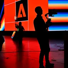 Sylwetki operatorów kamer na tle ekranu z logo Adobe. Czerwona podłoga odbija światła sceny.