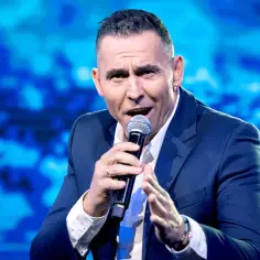 Mężczyzna w garniturze z mikrofonem, być może opowiada o tym, dlaczego Andrzej Kozłowski odszedł z kabaretu Pod Wyrwigroszem.