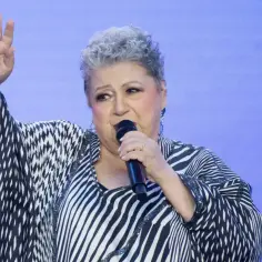 Ewa Bem z mikrofonem, w charakterystycznej, wzorzystej kreacji, wznosi rękę, jakby śpiewając swój przebój.